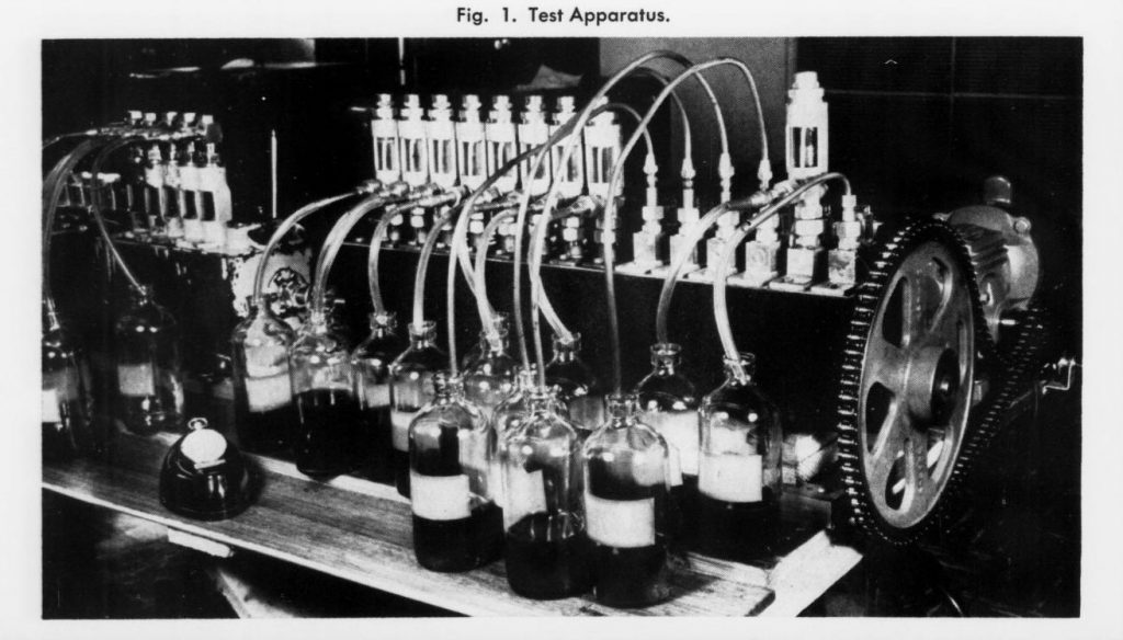 Scientific illustration in the form of a black-and-white photo of a device used to test how an engine uses up lubrication oil, consisting of a long shaft ending in a gear, lined with rows of tubes and nozzles, with a row of small bottles along the front.