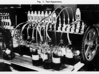 Scientific illustration in the form of a black-and-white photo of a device used to test how an engine uses up lubrication oil, consisting of a long shaft ending in a gear, lined with rows of tubes and nozzles, with a row of small bottles along the front.