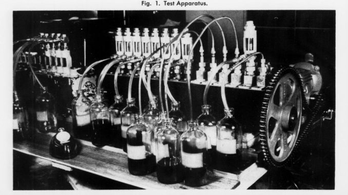 Scientific illustration in the form of a black-and-white photo of a device used to test how an engine uses up lubrication oil, consisting of a long shaft ending in a gear, lined with rows of tubes and nozzles, with a row of small bottles along the front.