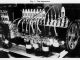Scientific illustration in the form of a black-and-white photo of a device used to test how an engine uses up lubrication oil, consisting of a long shaft ending in a gear, lined with rows of tubes and nozzles, with a row of small bottles along the front.