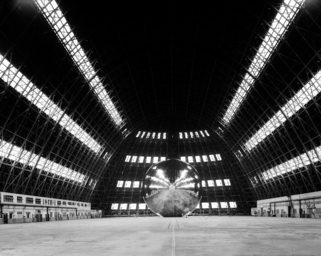 Scientific illustration in the form of a dramatic black and white photograph of a highly reflective sphere in the middle of a vast hangar lit by horizontal rows of lights so long, they seem to radiate out from a point of convergence somewhere behind the inflatable spacecraft.