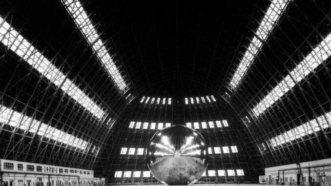 Scientific illustration in the form of a dramatic black and white photograph of a highly reflective sphere in the middle of a vast hangar lit by horizontal rows of lights so long, they seem to radiate out from a point of convergence somewhere behind the inflatable spacecraft.