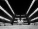 Scientific illustration in the form of a dramatic black and white photograph of a highly reflective sphere in the middle of a vast hangar lit by horizontal rows of lights so long, they seem to radiate out from a point of convergence somewhere behind the inflatable spacecraft.