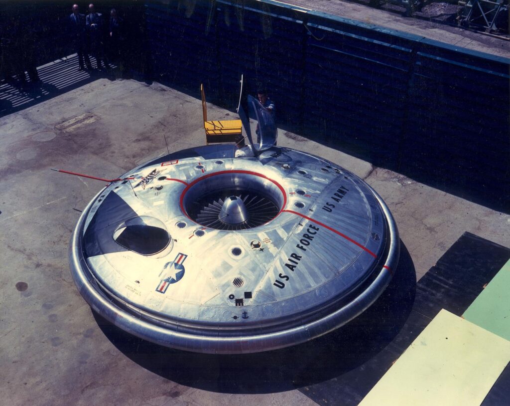 Scientific illustration of an Avrocar, a silver, disc-shaped aerial vehicle, gleaming steel and black vents, parked outside a shaded hangar.