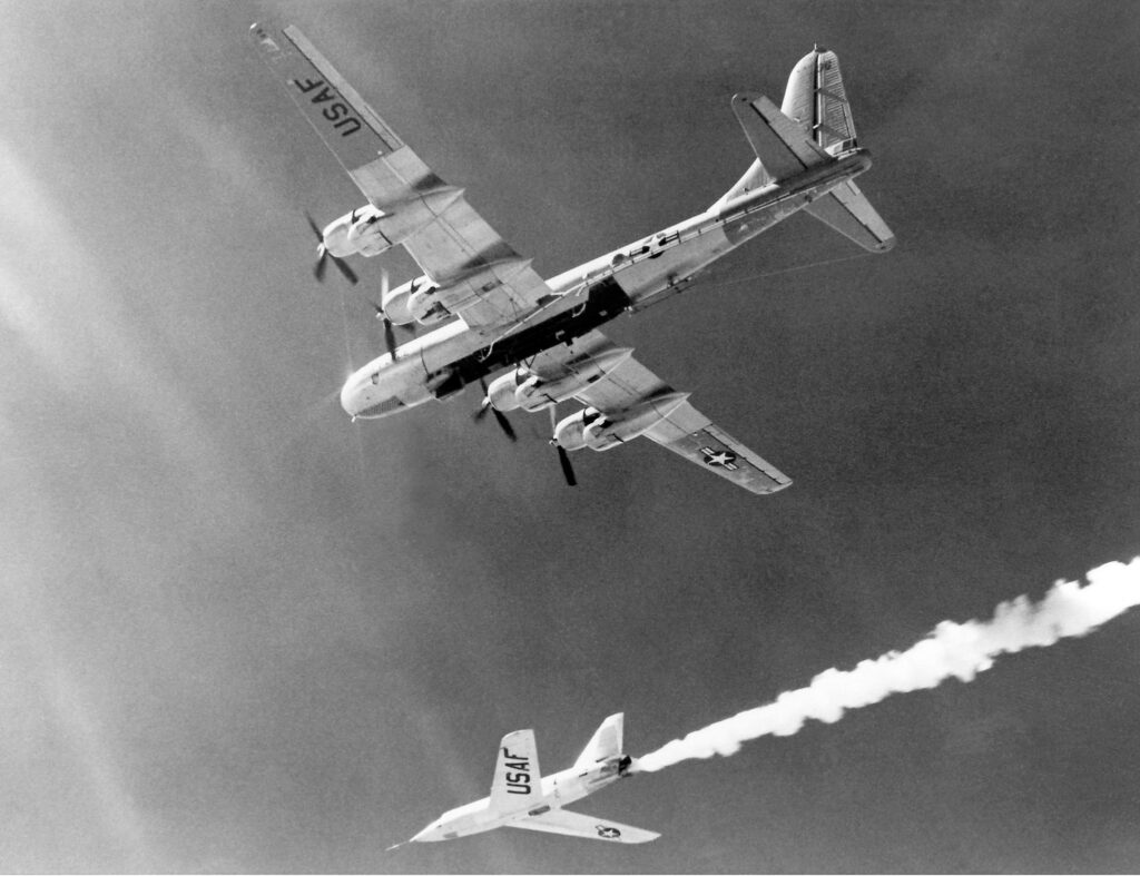 Scientific illustration of a supersonic rocket plane being launched from a bomber, a black-and-white photo of a shining steel dart leaving a trail of cloud beneath the massive shadow of its mothership.