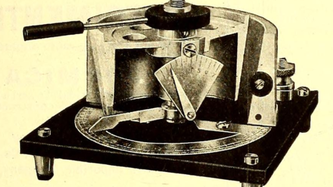 Scientific illustration of an electronic object that looks a little licke a pressure cooker with a cutaway side and some sort of an inditcator needle on the front. It's resting on a square stand with four tiny legs.