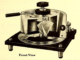 Scientific illustration of an electronic object that looks a little licke a pressure cooker with a cutaway side and some sort of an inditcator needle on the front. It's resting on a square stand with four tiny legs.