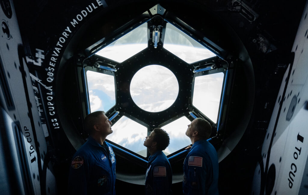 Scientific illustration of astronauts inside a replica of the International Space Station, a hexagonal window looking down on the Earth.