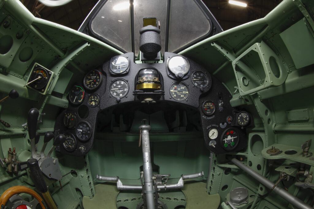 Scientific illustration, or photograph, really, of the green-and-black geometry of a WWII fighter plane cockpit, with an angled canopy above and a steering yoke in center of a small array of dials and indicators.