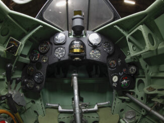 Scientific illustration, or photograph, really, of the green-and-black geometry of a WWII fighter plane cockpit, with an angled canopy above and a steering yoke in center of a small array of dials and indicators.