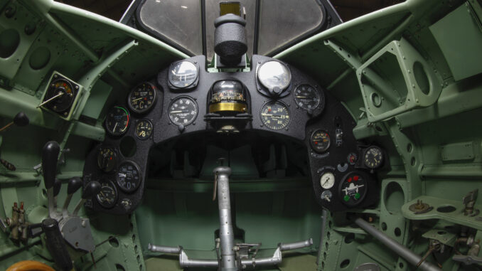 Scientific illustration, or photograph, really, of the green-and-black geometry of a WWII fighter plane cockpit, with an angled canopy above and a steering yoke in center of a small array of dials and indicators.