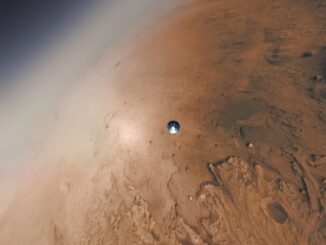 Scientific illustration in the form of an amplified photograph. The frame is filled mostly - but not entirely - with the red surface of Mars, a corner of deep space visible to the top left. In the center, barely visible, is a gleaming steel circle, a tiny droplet of metal descending to the rust-colored surface.