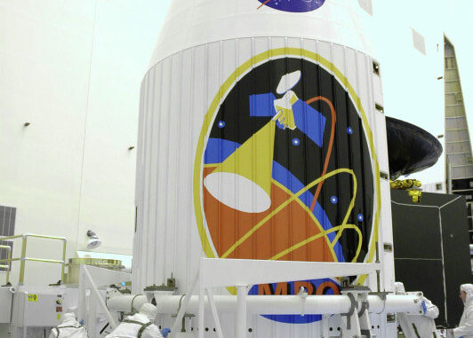 Scientific illustration: A crew in special suits executes s mechanical procedure with a large robot arm under an even larger painting of a mission emblem on the side of a space ship emblazoned with the NASA logo.