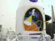 Scientific illustration: A crew in special suits executes s mechanical procedure with a large robot arm under an even larger painting of a mission emblem on the side of a space ship emblazoned with the NASA logo.
