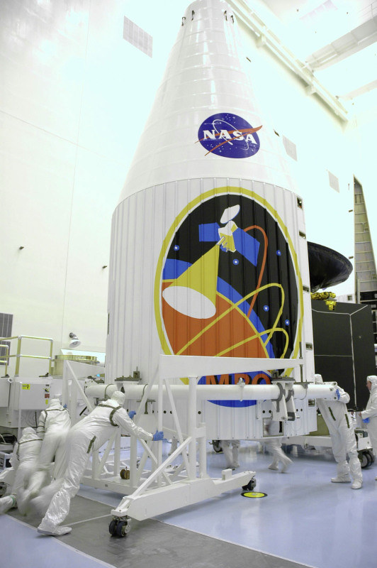 Scientific illustration: A crew in special suits executes s mechanical procedure with a large robot arm under an even larger painting of a mission emblem on the side of a space ship emblazoned with the NASA logo.