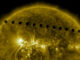 Scientific illustration of Venus transiting in front of the Sun, as captured by the Solar Dynamics Observetory satellite, a flaming metallic orb girdled by a series of perfectly circular silhouettes making a diagonal line from the upper left to the middle right of the image.