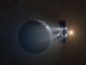 A dark spacecraft firing four small, glowing engines nears a round, rocky planet or planetoid. Perhaps it is Earth. The sun is small in the distance.
